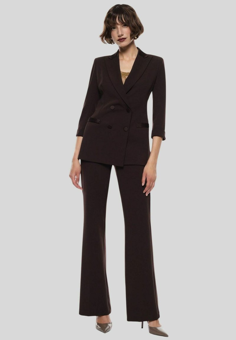 Abito marrone scuro con giacca doppiopetto su misura a tre quarti, abbinato a pantaloni a gamba larga e tacchi a punta.