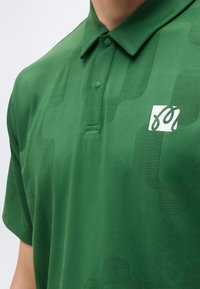 Polo vert avec des motifs géométriques subtils et un petit logo abstrait blanc sur le côté gauche de la poitrine, doté d'un col boutonné.