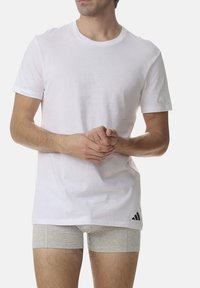 White cotton T-shirt with a crew neckline and short sleeves, featuring a black Adidas logo accent on the lower left side.