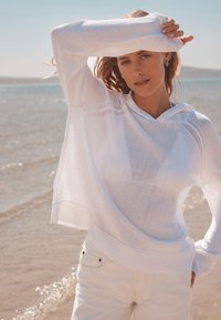 Sweat à capuche blanc léger avec une coupe ample, doté d'un ourlet et de poignets côtelés. Assorti à un jean clair, sur fond de plage.