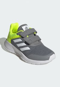 Chaussure de sport grise avec des accents jaunes, tige en maille, bandes Velcro, trois rayures blanches et une semelle en caoutchouc blanche. Surface texturée.