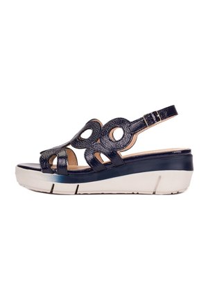 Sandale compensée en cuir verni noir avec découpes circulaires, bride de cheville ajustable et semelle épaisse blanche avec fond cranté.