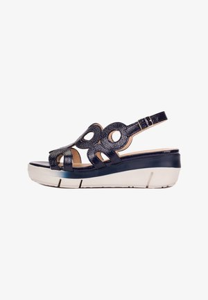 Sandale compensée en cuir verni noir avec découpes circulaires, bride de cheville ajustable et semelle épaisse blanche avec fond cranté.