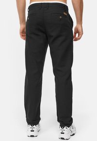 Black pants with a straight fit, made from a textured fabric, featuring two back pockets with buttons and a logo patch at the waistband.