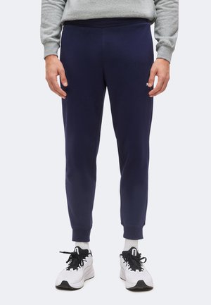 Persoon met een marineblauwe joggingbroek, een grijze trui, witte sokken en witte sneakers, staand tegen een effen lichte achtergrond.
