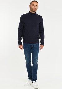 Maglione a collo alto blu navy realizzato in morbido tessuto a maglia, abbinato a jeans slim-fit e sneakers bianche. Design semplice con polsini e orlo a coste.