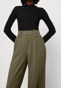 Schwarzes Langarmshirt kombiniert mit hoch taillierten, olivgrünen Hosen mit Falten und zwei großen runden Knöpfen an der Taille.
