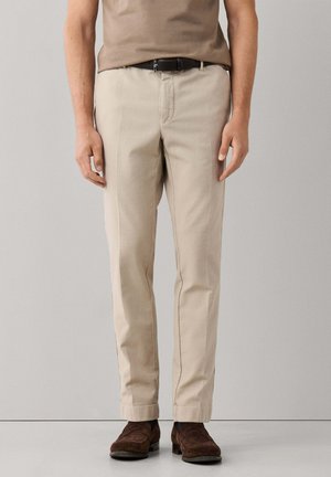 Man draagt beige rechte broek, bruine suède schoenen, zwarte riem en een bruin overhemd ingestopt, tegen een effen achtergrond.