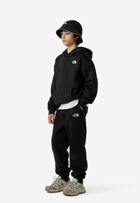 Svart hettegenser og joggebukse med brodert logo; kombinert med en svart bøttehatt og beige joggesko med grå detaljer.