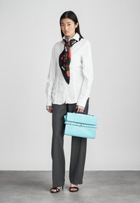 Vrouw in een witte gerimpelde blouse, zwarte bloemen sjaal, donkergrijze broek, zwarte open teen hakken, met een lichtblauwe gestructureerde handtas tegen een grijze achtergrond.