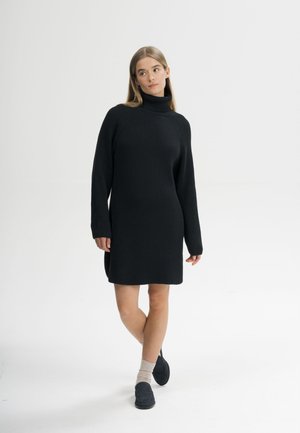 Frau in schwarzem geripptem Rollkragenkleid, beigen Socken und schwarzen Schuhen, stehend mit gekreuzten Beinen vor einem schlichten weißen Hintergrund.