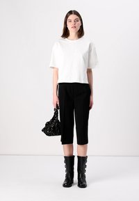 Tricou alb scurt cu maneci scurte, pantaloni negri cropped, botine negre până la genunchi cu catarame, ținând o geantă neagră cu model.