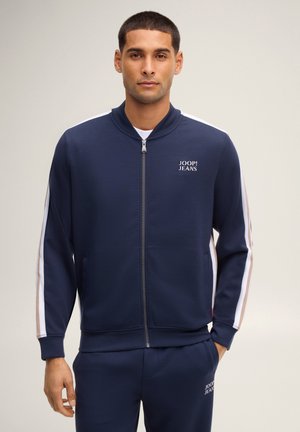 Man draagt een donkerblauw Joop! Jeans trainingspak met witte en beige strepen langs de mouwen en een logo op de linkerborst en broek.