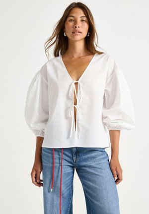 Blusa in lino bianco con scollatura a V profonda e dettaglio con fiocco davanti. Caratterizzata da maniche a sbuffo e una vestibilità rilassata. Abbinata a jeans a gamba larga blu.