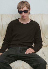 Junge Person mit hellbraunem Haar, die dunkle Sonnenbrille, einen schwarzen Pullover und schwarze Jeans trägt, sitzt auf einem cremefarbenen Ledersofa.