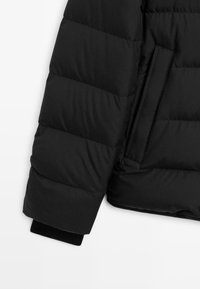 Puffy black jacket with a textured, quilted pattern. Features a side pocket and a contrasting black cuff at the sleeve opening.