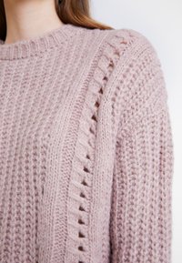 Pull tricoté rose pâle avec des motifs en torsades côtelées et ajourées sur l'épaule et la poitrine, porté par une personne aux longs cheveux bruns.