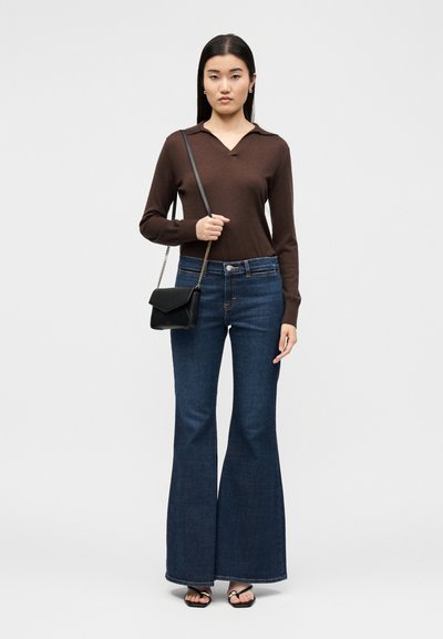 Γυναίκα που φοράει καφέ μακρυμάνικη μπλούζα, σκούρο μπλε καμπάνα τζιν, μαύρα σανδάλια και κρατάει μια μικρή μαύρη τσάντα χιαστί.