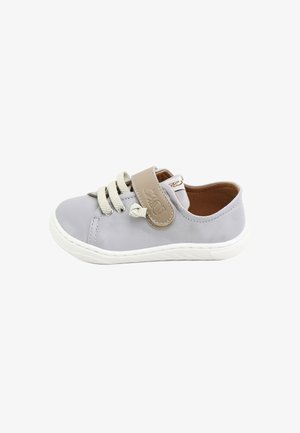 Zapatilla deportiva infantil de color gris claro con suela blanca, tira de velcro beige y cordones crema sobre fondo blanco.