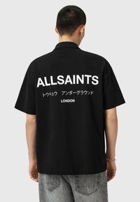Juoda trumpa rankovėmis marškinėliai su apykakle, su dideliu baltu užrašu "ALLSAINTS" ir japoniškais rašmenimis ant nugaros, pagaminti iš medvilnės.