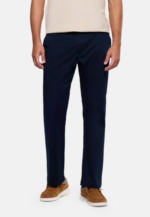 Hombre vestido con pantalones azul marino de pierna recta, camiseta beige, calcetines grises y zapatos mocasines marrones, de pie frente a un fondo blanco.