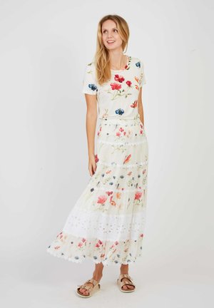 Kobieta z długimi blond włosami, ubrana w biały kwiecisty krótki top i pasującą długą kwiecistą spódnicę oraz beżowe sandały, stojąca na białym tle.