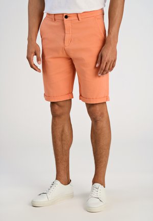 Persikofärgade shorts med slät framsida, uppsvängda fållar och ett enda knappstängning. Bärs med vita sneakers på en neutral bakgrund.