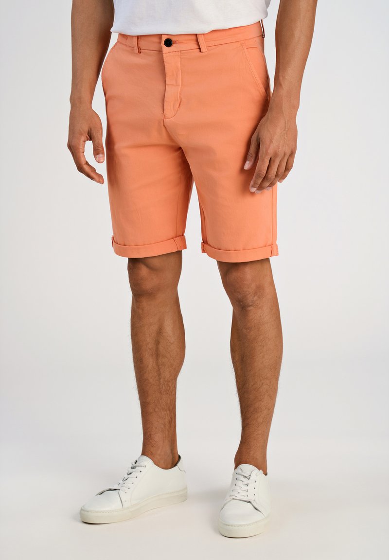 Shorts de couleur pêche avec une taille plate, des ourlets retournés et une fermeture à un bouton. Portés avec des baskets blanches sur un fond neutre.