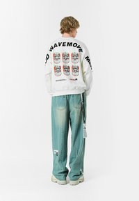 Sudadera blanca con estampado gráfico de seis latas de colores en la parte trasera, con gran texto negro que dice "WAVEMOVE". Jeans de corte holgado y color azul claro.