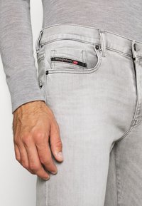 Unos vaqueros de denim gris claro presentan un diseño ligeramente desgastado. El bolsillo trasero lleva una etiqueta negra con texto rojo, y se puede ver un cierre de cremallera.