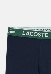 Boxer in cotone blu navy con cintura elastica verde decorata con il logo bianco "LACOSTE" e l'emblema del coccodrillo. Texture liscia, design aderente.