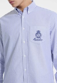 Camisa de botón a rayas en azul claro y blanco con un bolsillo en el pecho que presenta un detalle de logo bordado. Fabricada en tejido entrelazado, de corte clásico.