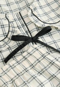 Placket in ovratnik iz lahkega karirastega blaga v krem in mornarsko modri, z črnimi robovi in privezano pentljo. Teksturne ruffle na robovih.