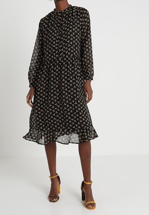 Kobieta w czarnej, midi sukience z długim rękawem, ozdobionej drobnym kwiatowym wzorem i falbaną u dołu, w połączeniu z musztardowo-żółtymi sandałami na wysokim obcasie.