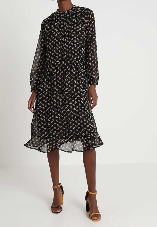 Femme portant une robe midi noire à manches longues, imprimé floral discret et ourlet à volants, associée à des sandales à talons hauts jaune moutarde.