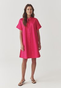 Robe chemise fuchsia avec un col haut, des manches courtes et une silhouette droite. Tissu lisse et fermeture à boutons centrée.