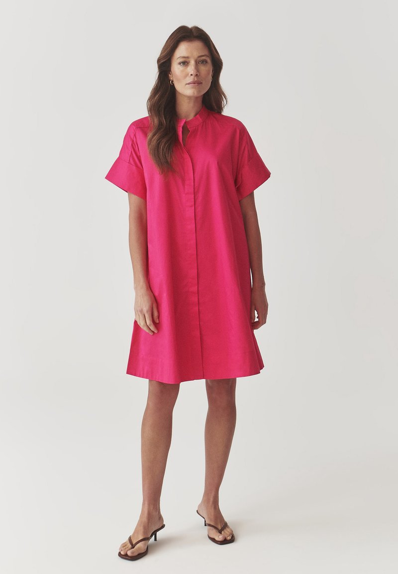Robe chemise fuchsia avec un col haut, des manches courtes et une silhouette droite. Tissu lisse et fermeture à boutons centrée.