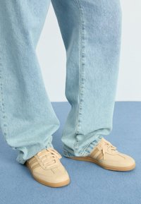 Beige lädersneakers med tre tunna bruna ränder, tillsammans med lösa ljusblå denimjeans. Slät yta, rund tå, synliga snören.