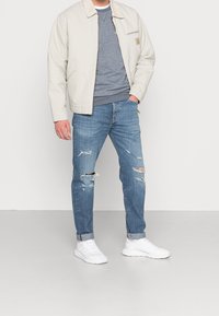 Hombre con chaqueta beige, suéter gris, jeans azules desgastados y zapatillas blancas, de pie contra un fondo liso con la mano en el bolsillo.
