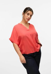 Blusa coral con escote en V, de manga corta, corte holgado y textura suave. Llevada con jeans azul oscuro de corte recto clásico.