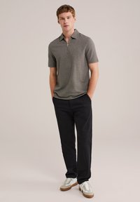 Camisa polo gris con cremallera y mangas cortas, combinada con pantalones negros y zapatillas blancas. La polo tiene una tela texturada y un corte relajado.