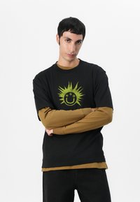 Black t-shirt with green sun graphic, layered over a brown long-sleeve shirt. Cotton material, relaxed fit, crew neckline.