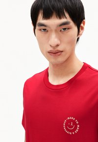 T-shirt en coton rouge avec un col rond et des manches courtes, arborant un logo circulaire de couleur claire et du texte sur le devant.