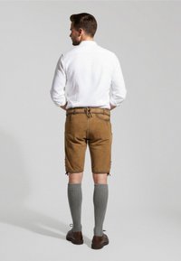 Brune lædershorts med sidelommer, båret med grå knælange sokker. Modellen har en hvid skjorte med knaplukning på, set fra ryggen.