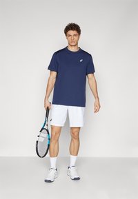Herren Sport-T-Shirt in Marineblau mit kurzen Ärmeln, kombiniert mit weißen Shorts. Hält einen Tennisschläger mit blauen Akzenten und trägt weiße Sneaker.