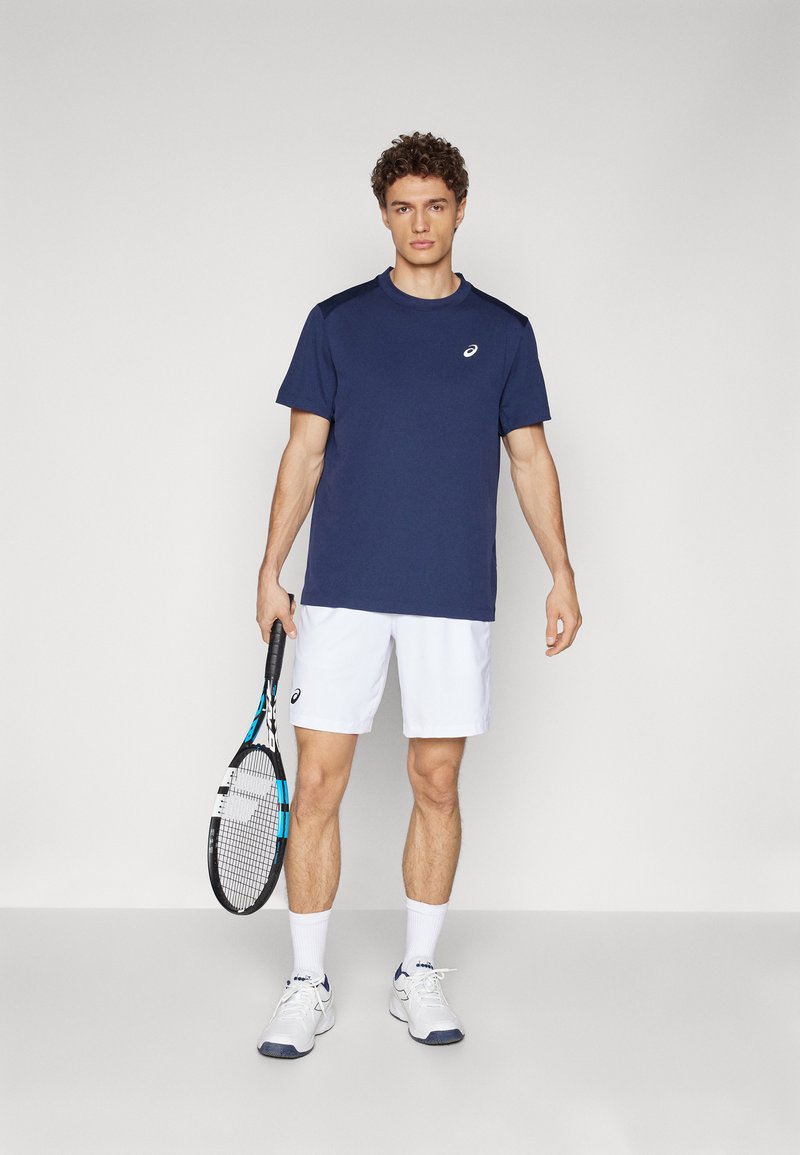 Herren Sport-T-Shirt in Marineblau mit kurzen Ärmeln, kombiniert mit weißen Shorts. Hält einen Tennisschläger mit blauen Akzenten und trägt weiße Sneaker.
