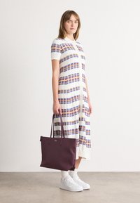 Γυναίκα που στέκεται σε ένα λευκό φόρεμα με μπλε και πορτοκαλί καρό ρίγες, λ�ευκά αθλητικά παπούτσια και κρατώντας μια μεγάλη σκούρα μωβ τσάντα.