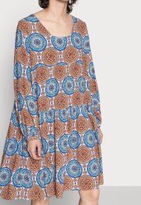 Vestido estampado con escote redondo, que presenta estampados circulares en azul, naranja y rojo sobre un fondo claro. Mangas largas y falda fruncida.