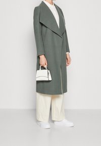 Lång, grön kappa med bred krage och öppen front, tillsammans med en vit skjorta, ljusbeiga byxor och en liten vit handväska. Vita sneakers.