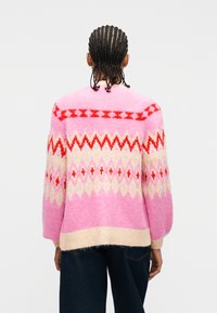 Άτομο με πλεξούδες φοράει πουλόβερ με ροζ και κόκκινο μοτίβο και σκούρο παντελόνι, εμφανιζόμενο από πίσω σε απλό φόντο.